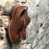 A tan vegan leather brogue dress shoe, lace up with 3 eyelets. Tan and dark brown sole with rubber grip for traction. Shown against a brick and concrete wall.